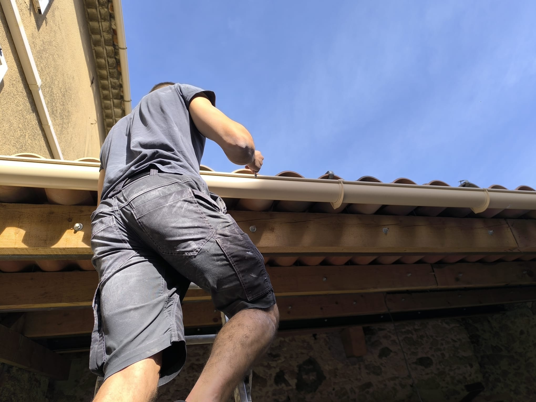 Artisan intervenant sur une gouttière en bordure de toiture, sous un ciel bleu